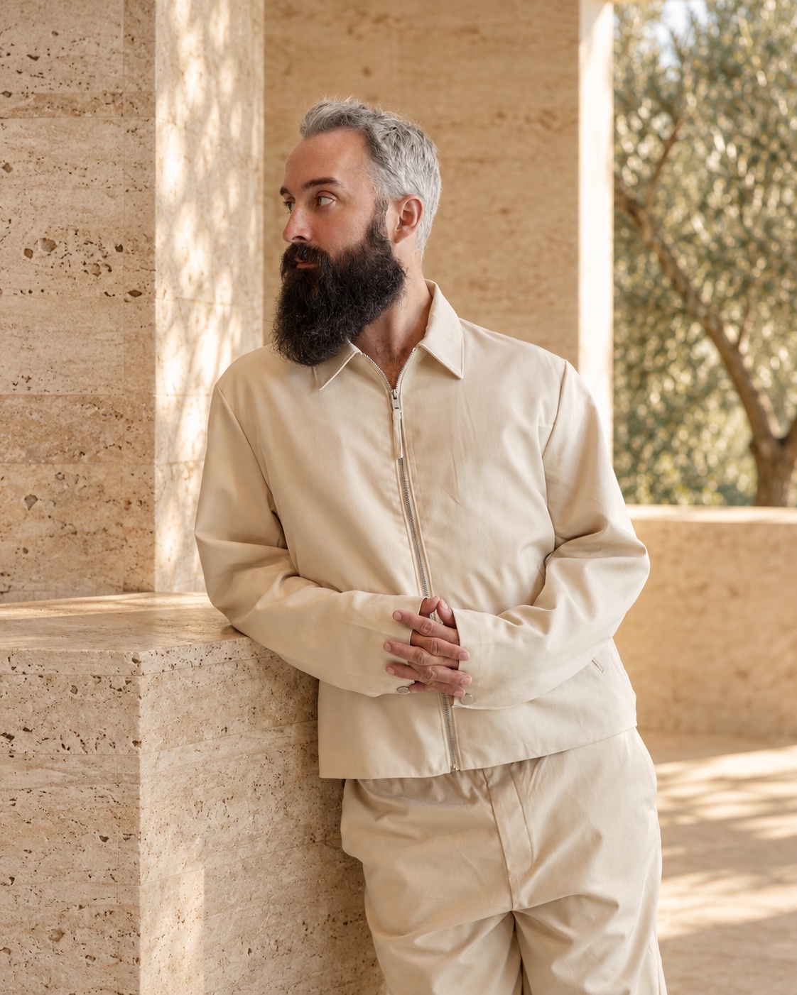 Dylan in a cream outfit, standing beside a sunlit stone column