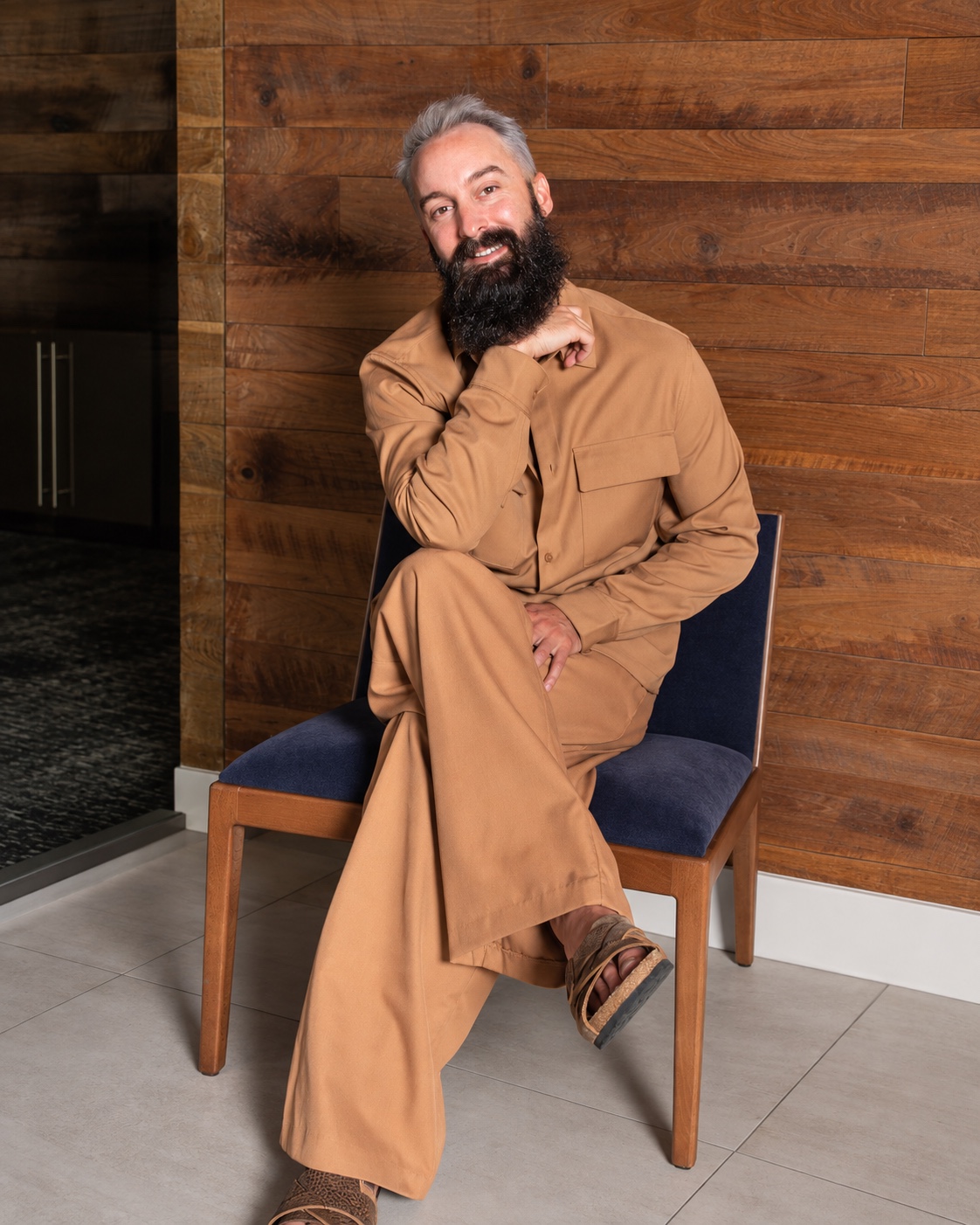 Dylan smiling in a tan outfit, seated against a wood-paneled wall